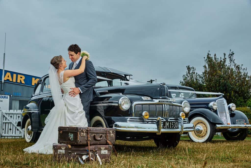 A bride and groom embrace in front of two vintage cars with stacked suitcases on the grass nearby, under a cloudy sky—a perfect moment captured by a Kaikōura wedding photographer at a New Zealand coastal wedding.